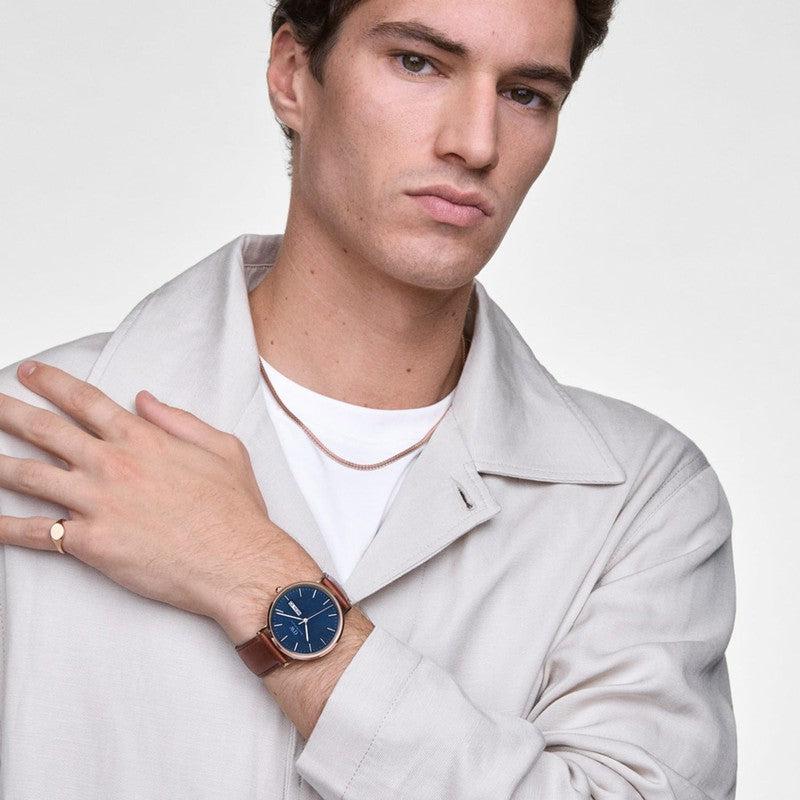 Daniel Wellington Classic watch with a blue dial, rose gold case, and brown leather St Mawes strap on a man's wrist.