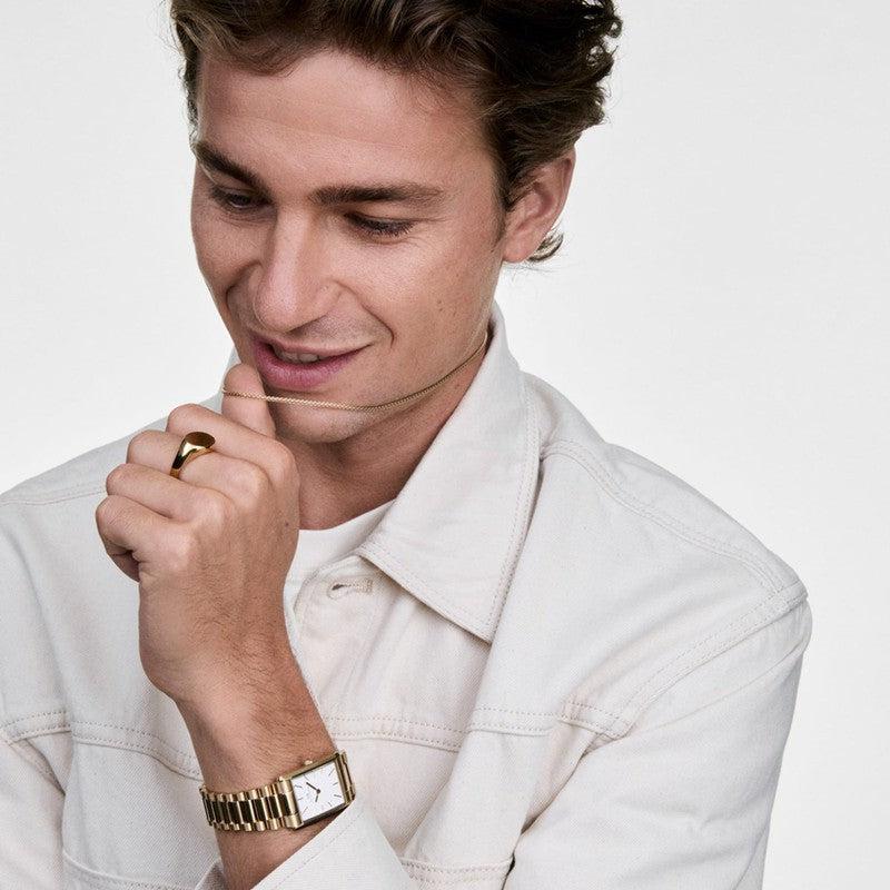 Unisex Daniel Wellington gold mesh necklace in stainless steel, worn with a matching gold watch and ring.